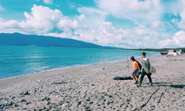 Due ragazzi passeggiano sulla spiaggia della Maremma Toscana, uno con una tavola da surf, mentre il mare azzurro lambisce la riva.