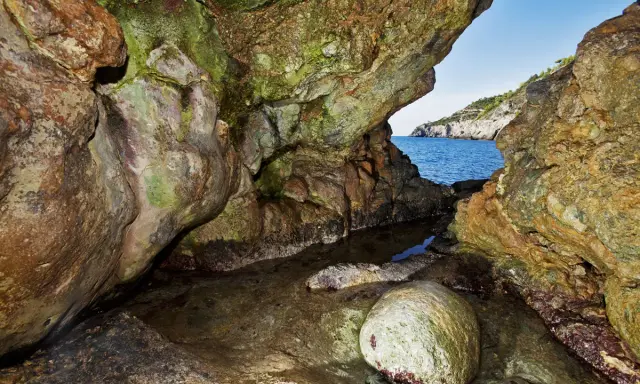 L'entrata di una grotta marina, la cala del bove, con rocce muschiose e un piccolo specchio d'acqua che riflette il blu del mare e il cielo.