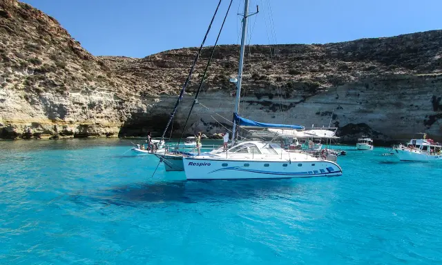 Catamarano "Respiro" in acque cristalline di un'isola sotto la Sicilia, con scogliere rocciose e altre barche.