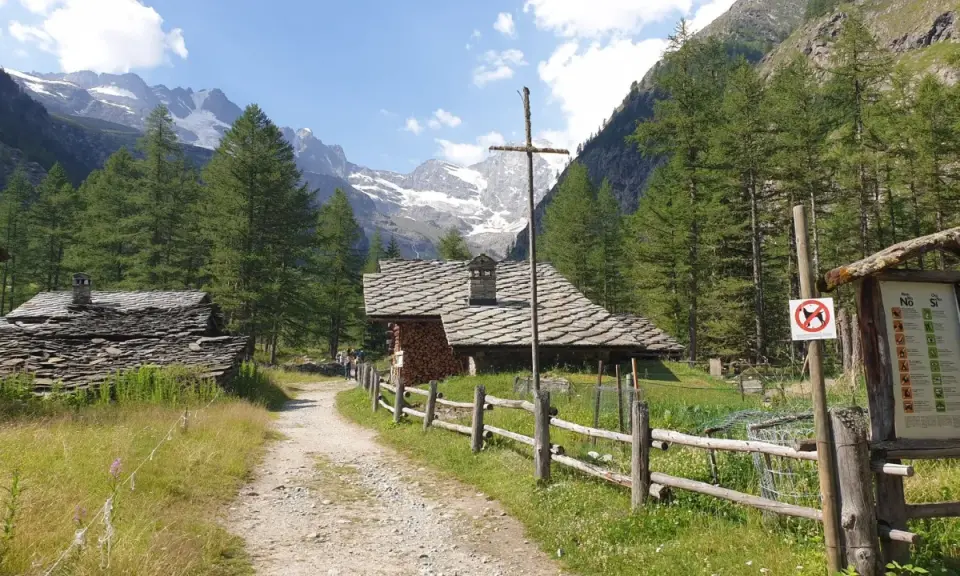 Sentiero di montagna con baite in pietra e tettoie, ideale per cogne escursioni facili. Montagne innevate sullo sfondo.