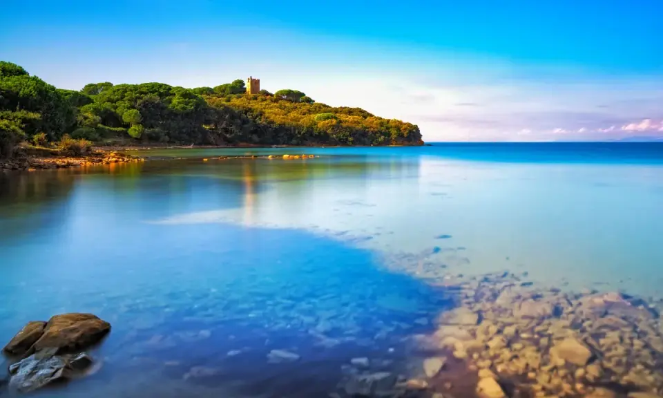 Torre medievale su una collina verdeggiante affacciata sul limpido **golfo di Follonica**. Acque calme e trasparenti riflettono il cielo azzurro.