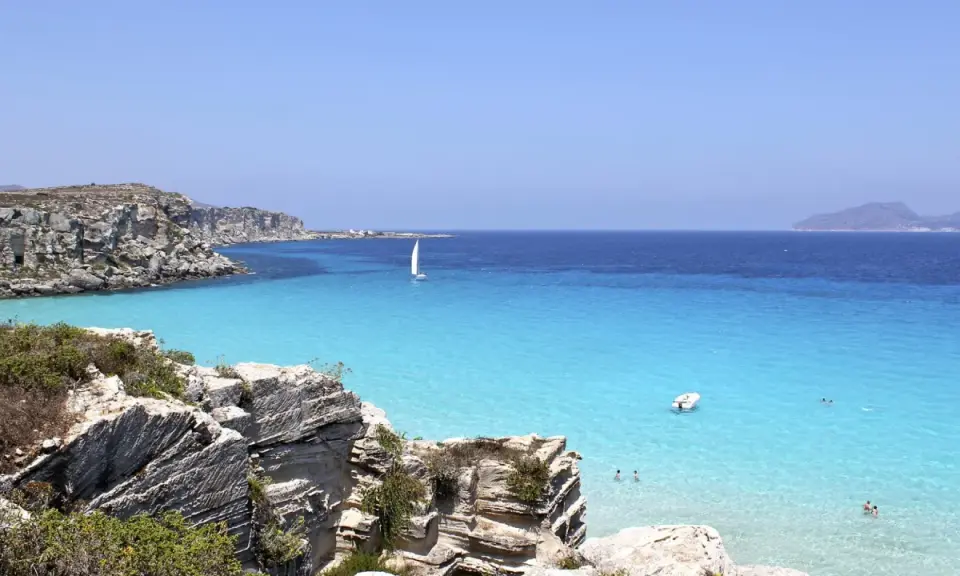 Scogliere rocciose e mare cristallino a Favignana. Un veliero solca le acque turchesi, perfetto per scoprire cosa vedere a Favignana in un giorno.