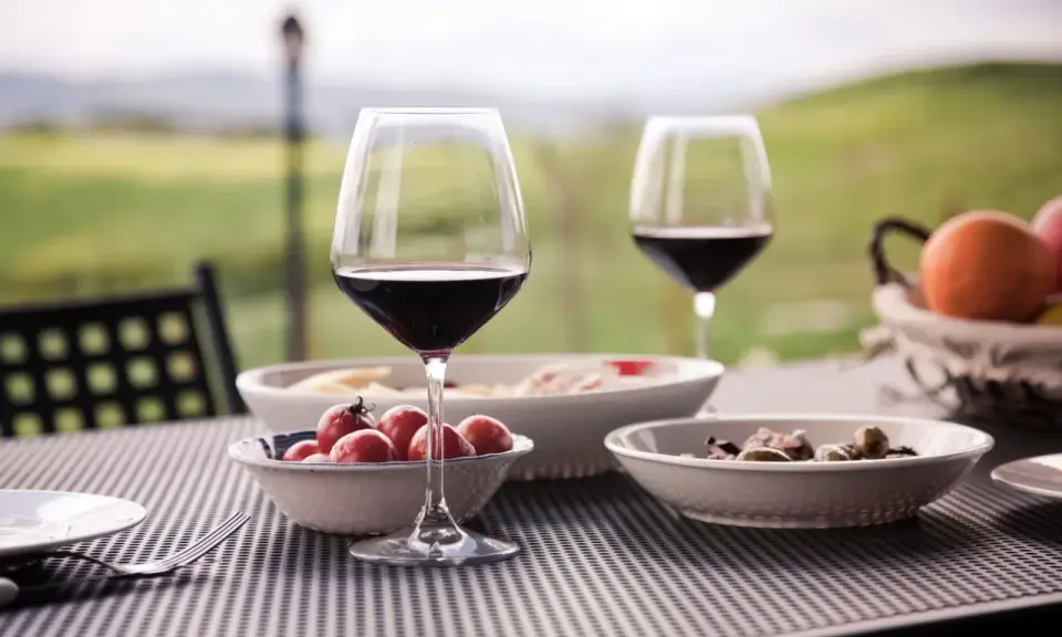 Un bicchiere di vino rosso e stuzzichini su un tavolo all'aperto, con un paesaggio collinare sullo sfondo, perfetto per un percorso enogastronomico toscana.