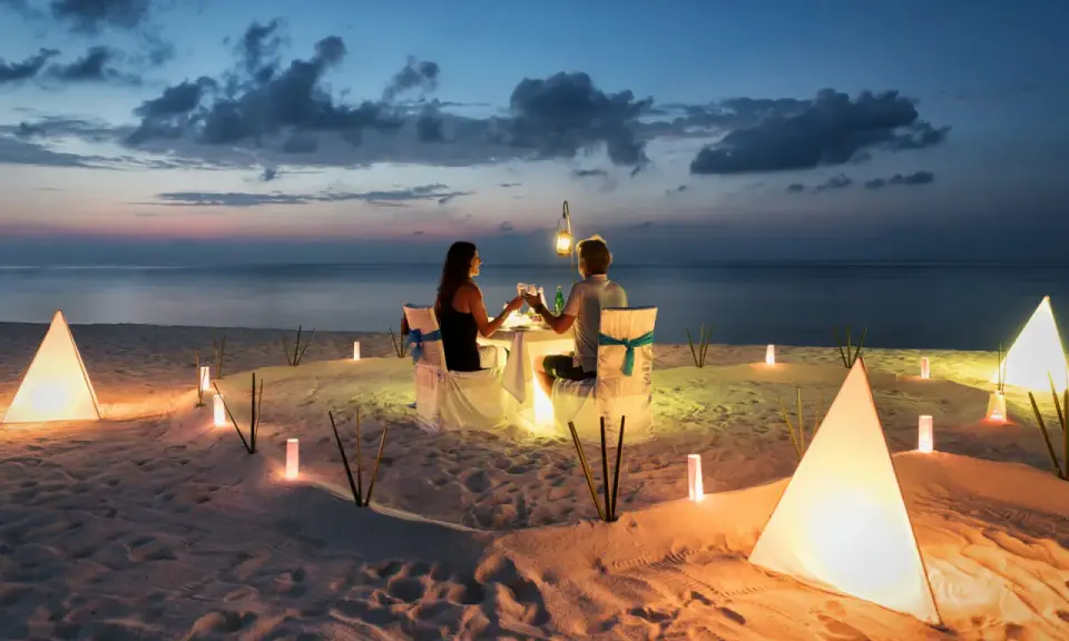 Una coppia al mare di sera, seduta a tavola per una cena romantica illuminata da lanterne e luci soffuse sulla sabbia.