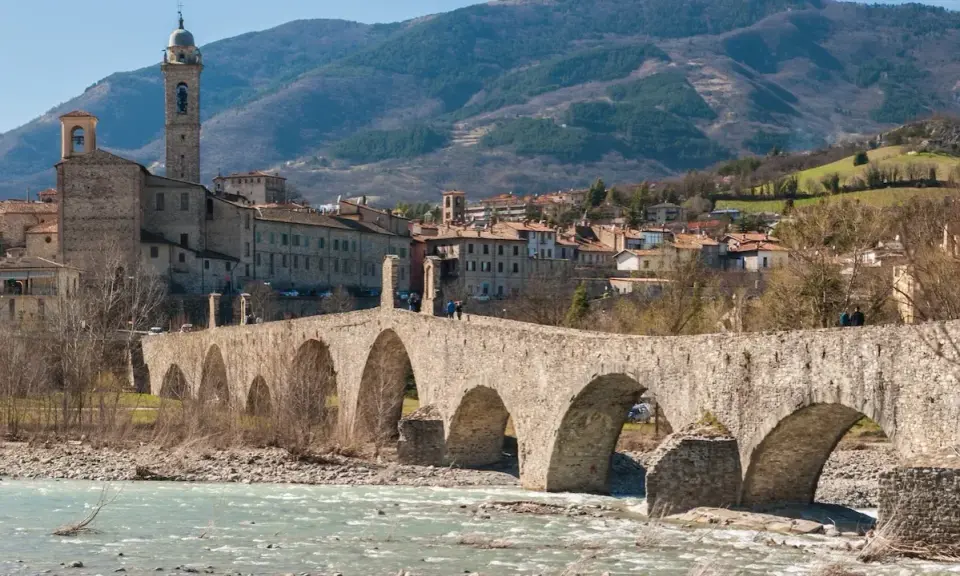 Ponte medievale in pietra attraversa un fiume, con un borgo storico e colline sullo sfondo. Un gioiello tra i borghi medievali dell'Emilia Romagna.