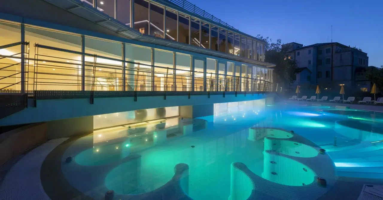 Piscina termale illuminata di notte, con vapori che salgono. Un edificio moderno con ampie vetrate si affaccia sull'acqua turchese, evocando un'atmosfera di relax e benessere, come in antiche terme preistoriche a Padova.