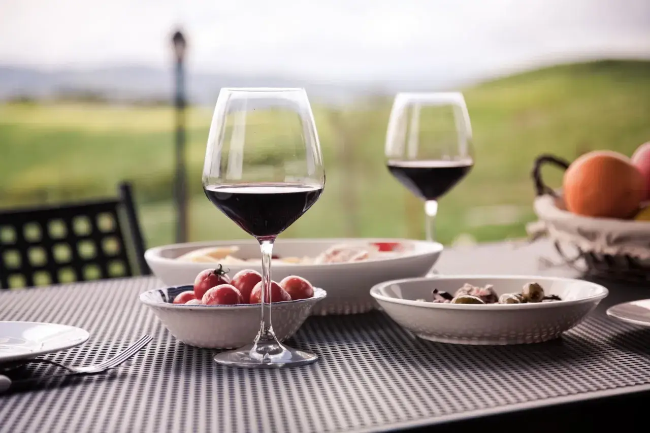 Un bicchiere di vino rosso e stuzzichini su un tavolo all'aperto, con un paesaggio collinare sullo sfondo, perfetto per un percorso enogastronomico toscana.