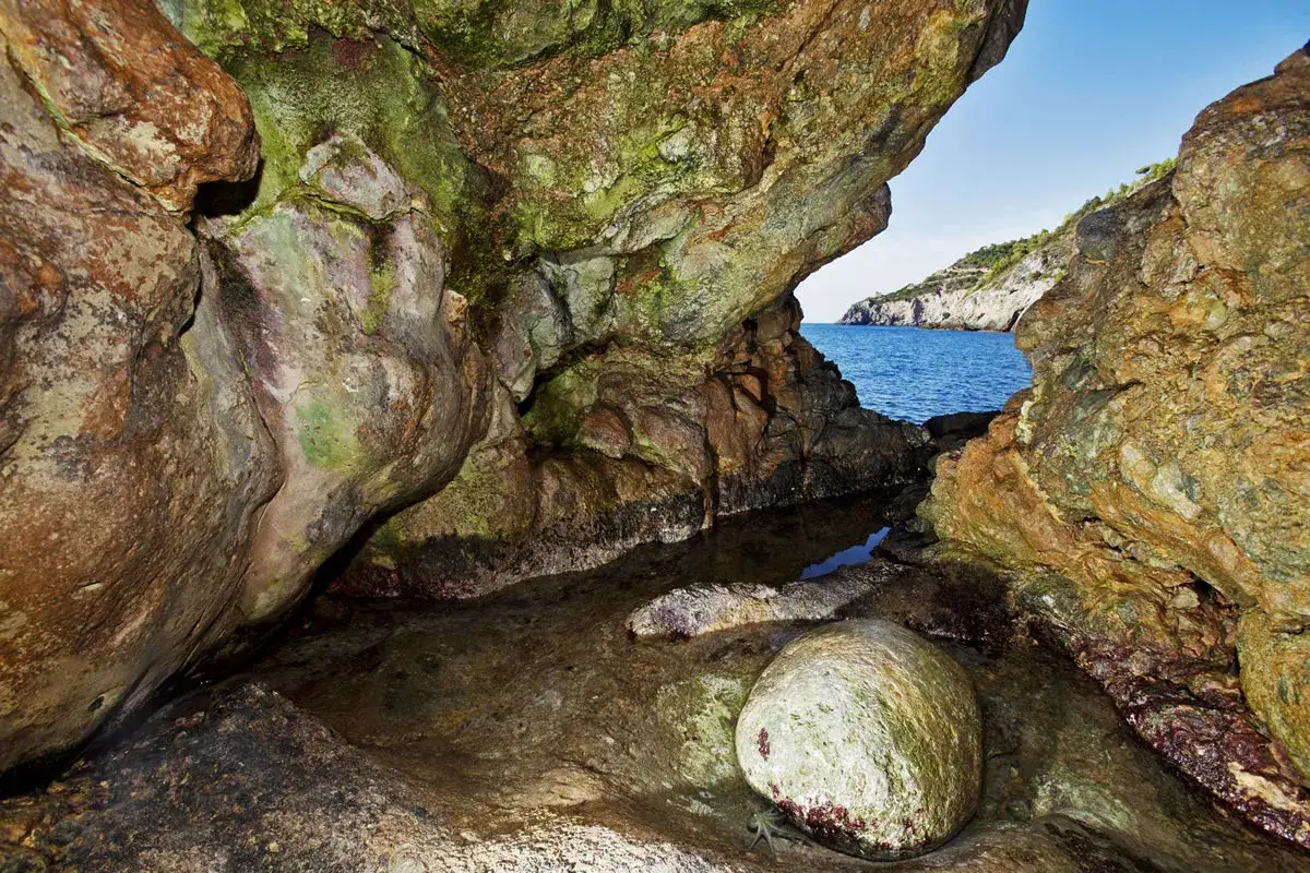 L'interno di una grotta marina, la Cala del Bove, con rocce muschiose e una pozza d'acqua che riflette il blu del mare e il cielo limpido.