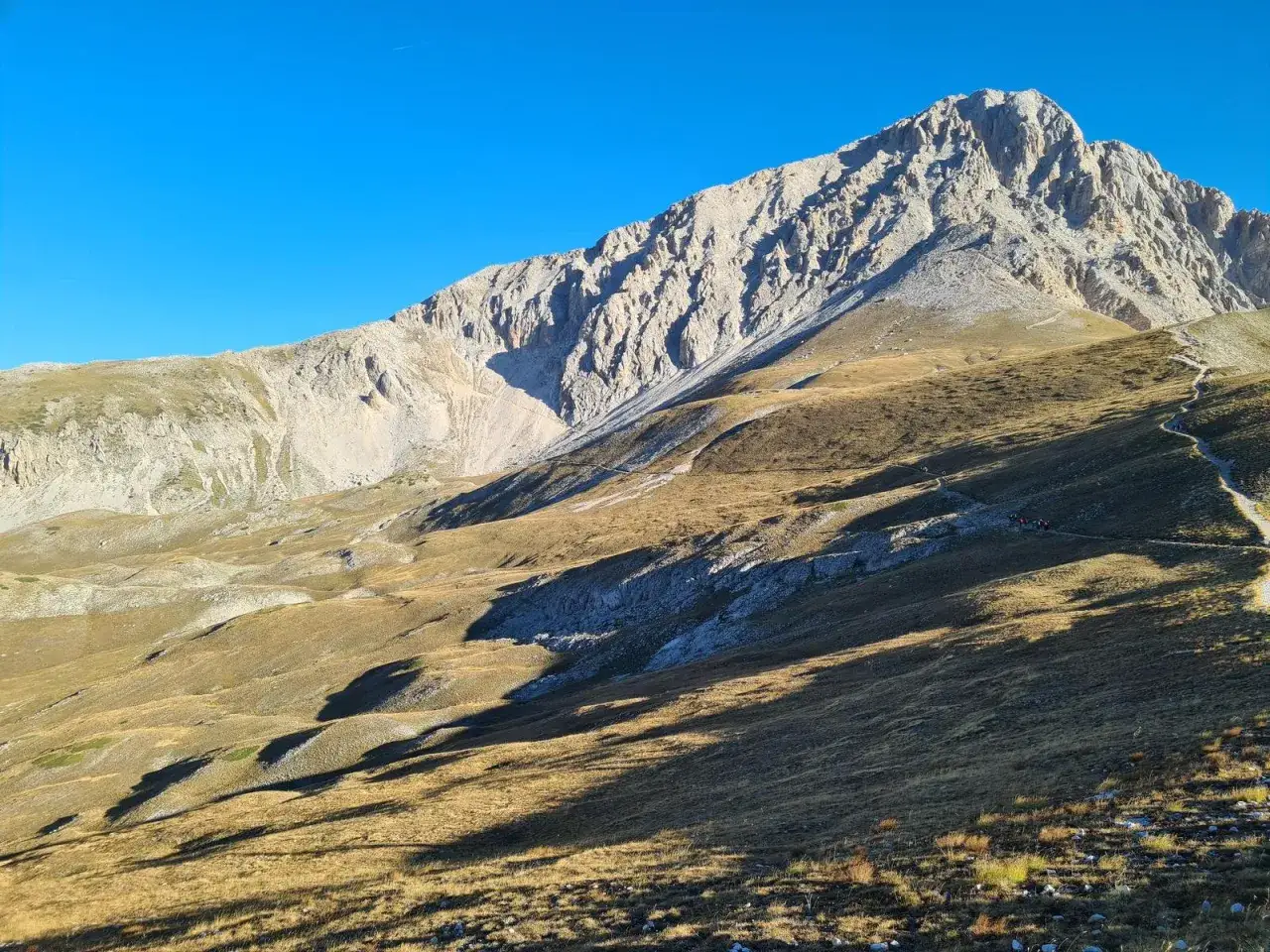 Sentiero tortuoso tra i prati dorati del Gran Sasso, dove piccoli alpinisti si avventurano verso la vetta sotto un cielo azzurro intenso.