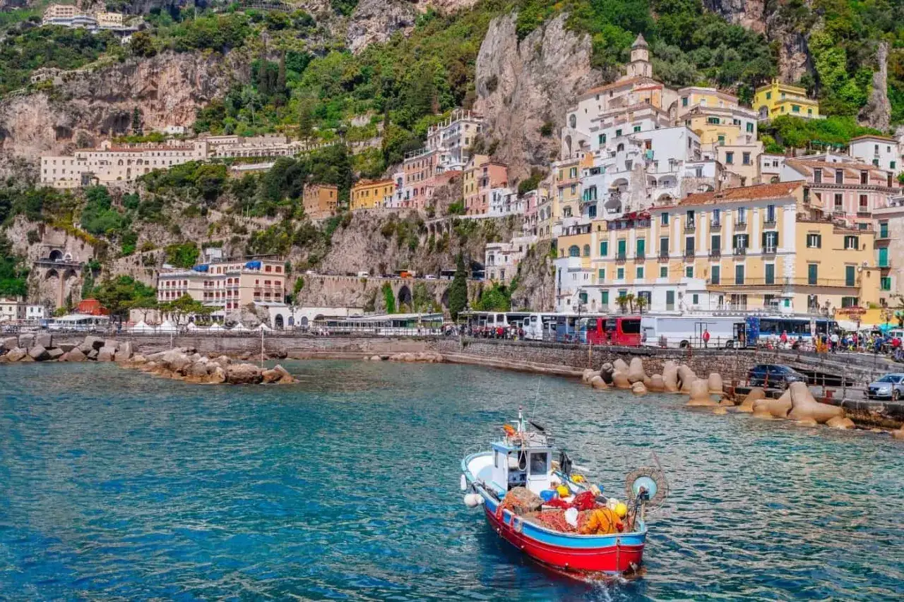 Un peschereccio colorato naviga nelle acque azzurre, con case a cascata sulla scogliera. Un'ispirazione per come visitare la Costiera Amalfitana.