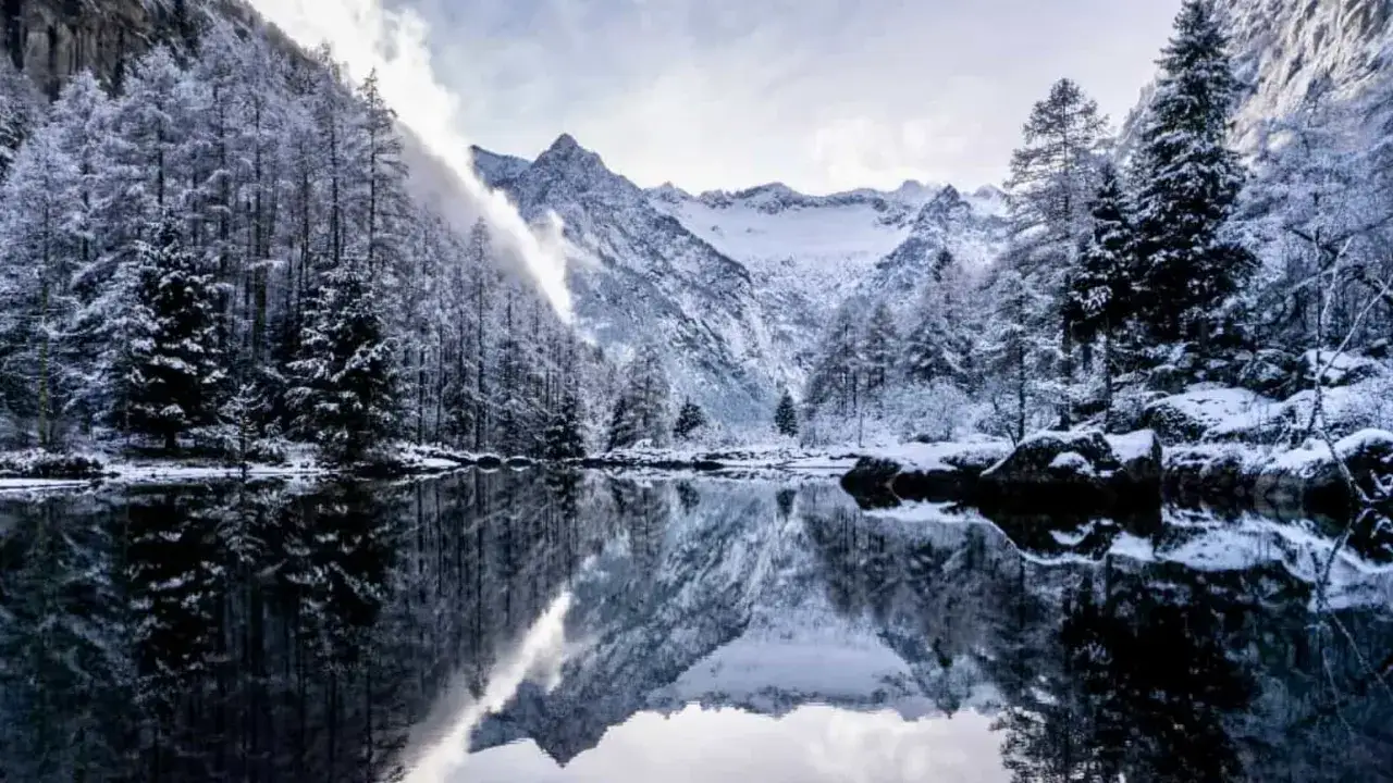 Val di Mello inverno: alberi innevati e montagne maestose si specchiano in un lago calmo, creando un paesaggio da fiaba.