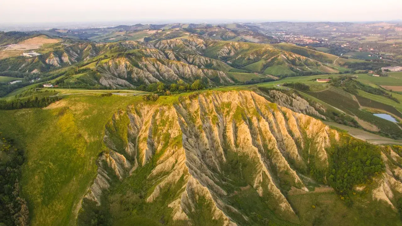 Dolci colline verdi e calanchi argillosi disegnano il paesaggio degli Appennini in Emilia Romagna, con un piccolo lago e case sparse.