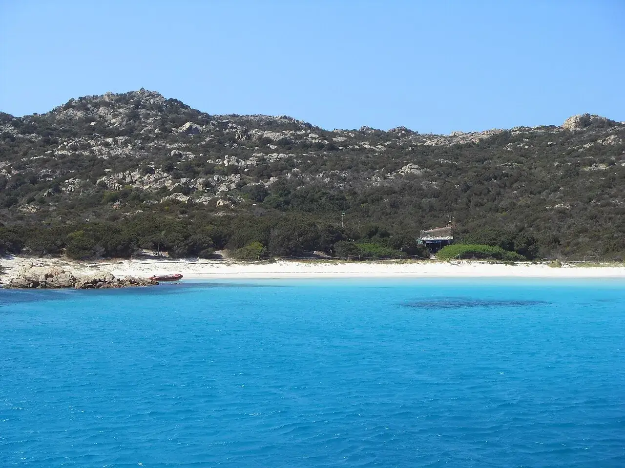 Acque cristalline e una spiaggia bianca sull'isola di Budelli, con una piccola casa e un gommone rosso sulla riva.