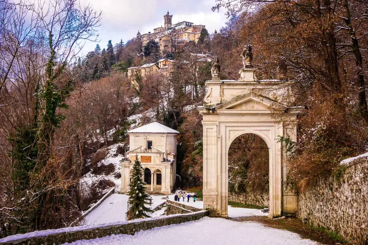 Un arco monumentale e un piccolo edificio religioso innevati, con un borgo arroccato sullo sfondo. Un luogo suggestivo per le cose da fare a Varese.