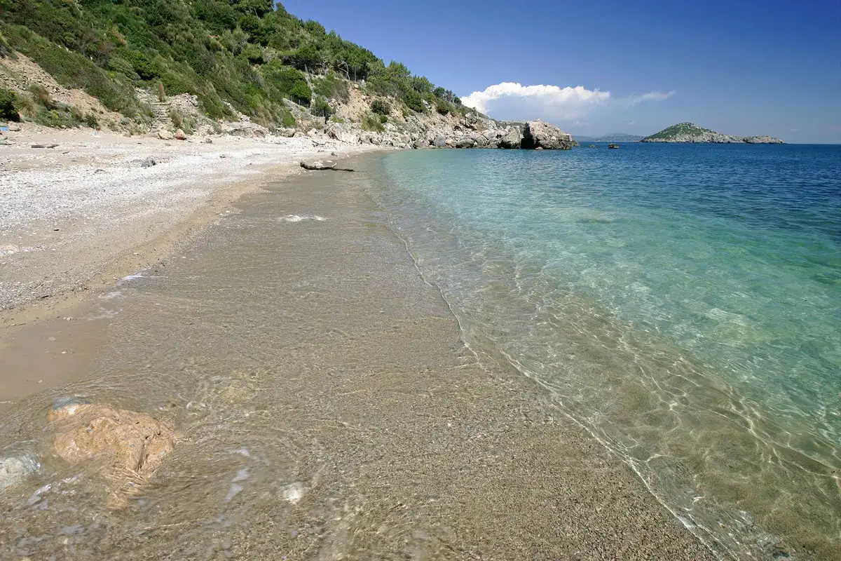 Spiaggia di ciottoli e sabbia con acqua cristallina, vista verso il mare aperto e un'isola in lontananza, vicino al porto di Ercole.