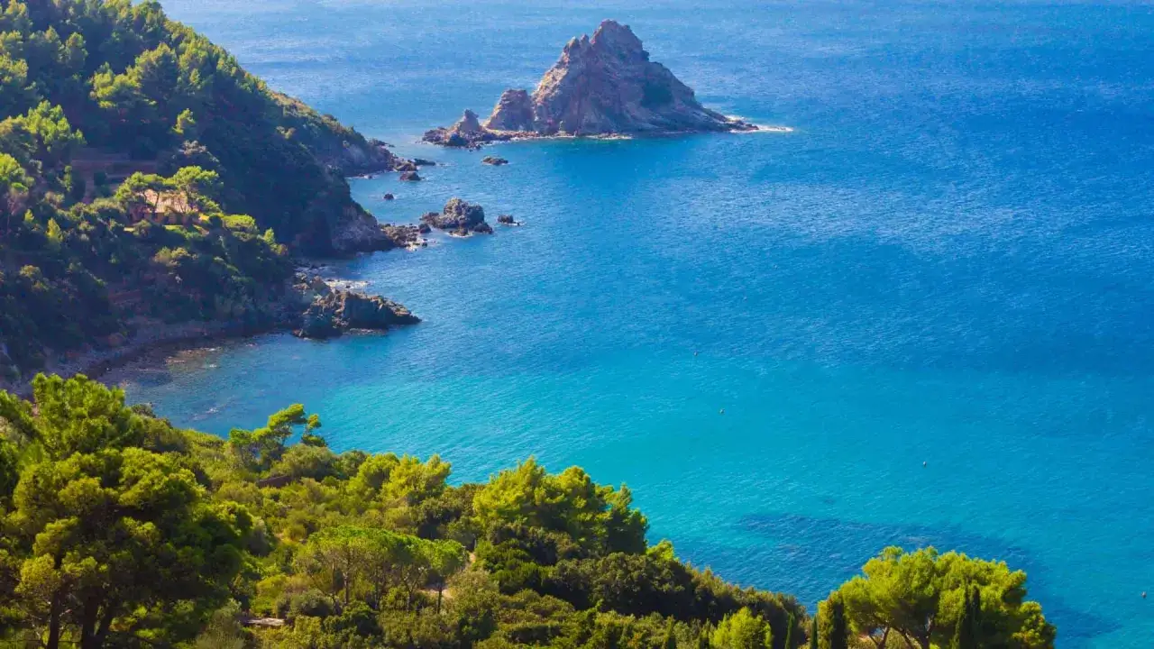 Scogliera verdeggiante e mare cristallino nella Maremma Toscana. Un'isola rocciosa emerge dalle acque turchesi.