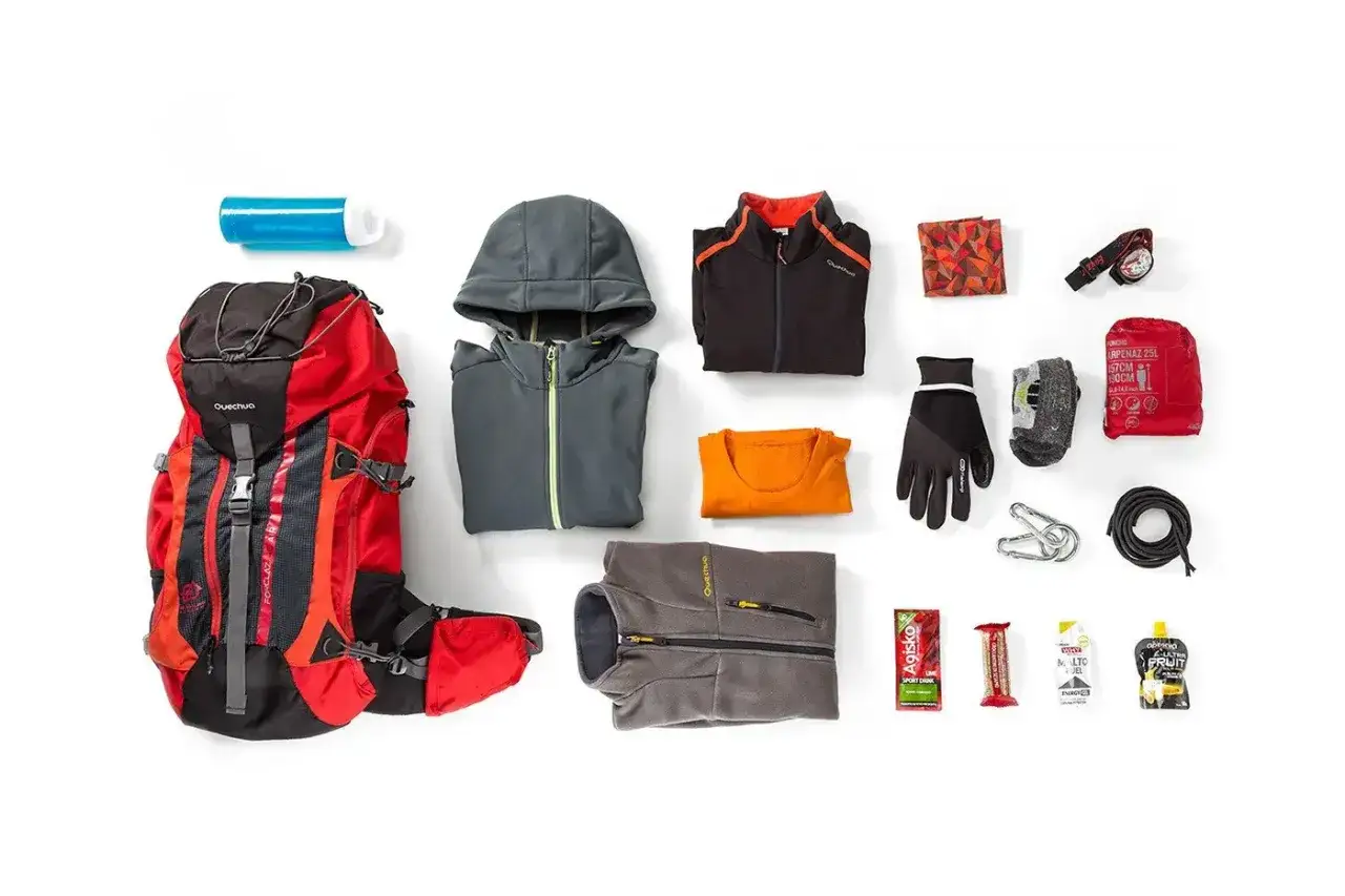 Zestaw do camminare in montagna: zaino rosso, pile, giacca, maglia, guanti, borraccia, kit di pronto soccorso, torcia, corda e snack energetici.