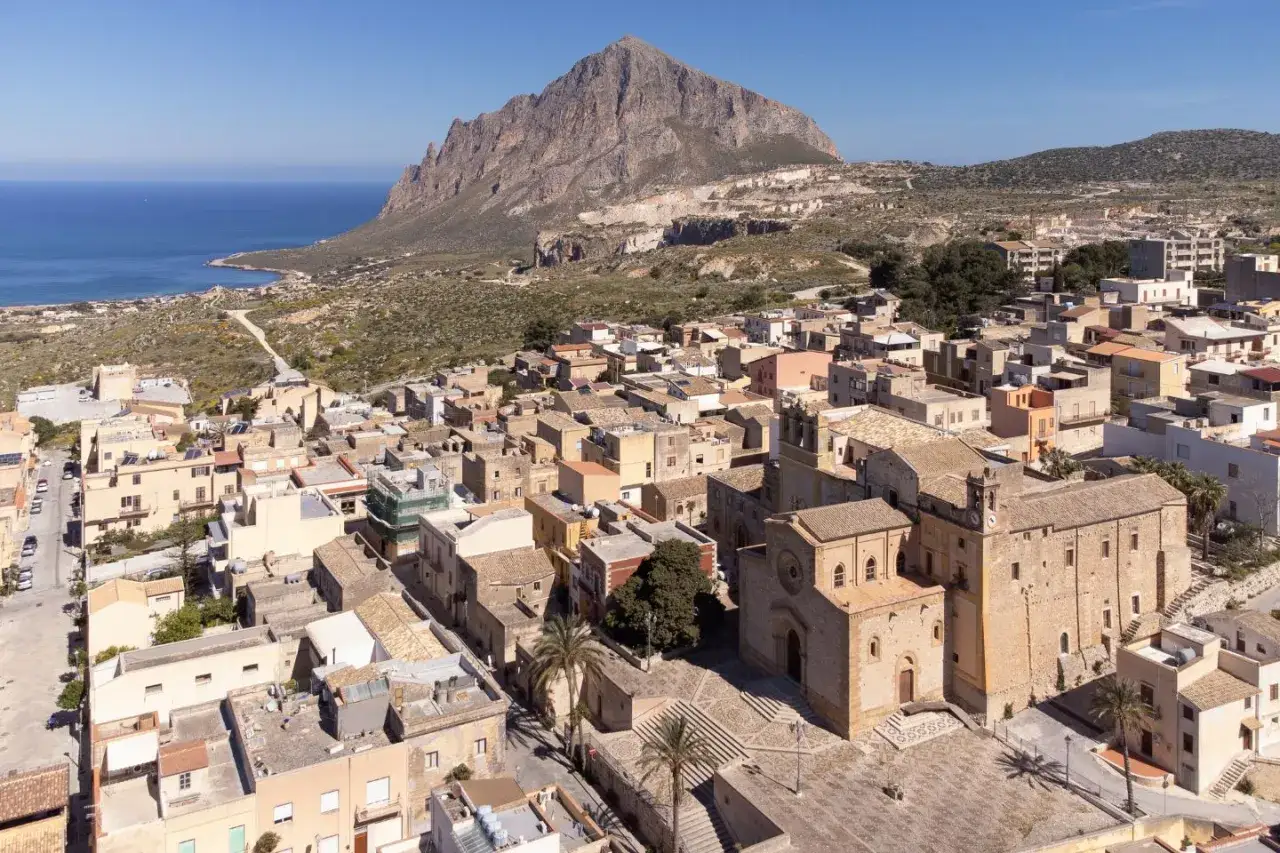 Un borgo da visitare in Sicilia, con case color pastello e una chiesa imponente, incastonato tra il mare blu e una montagna imponente.