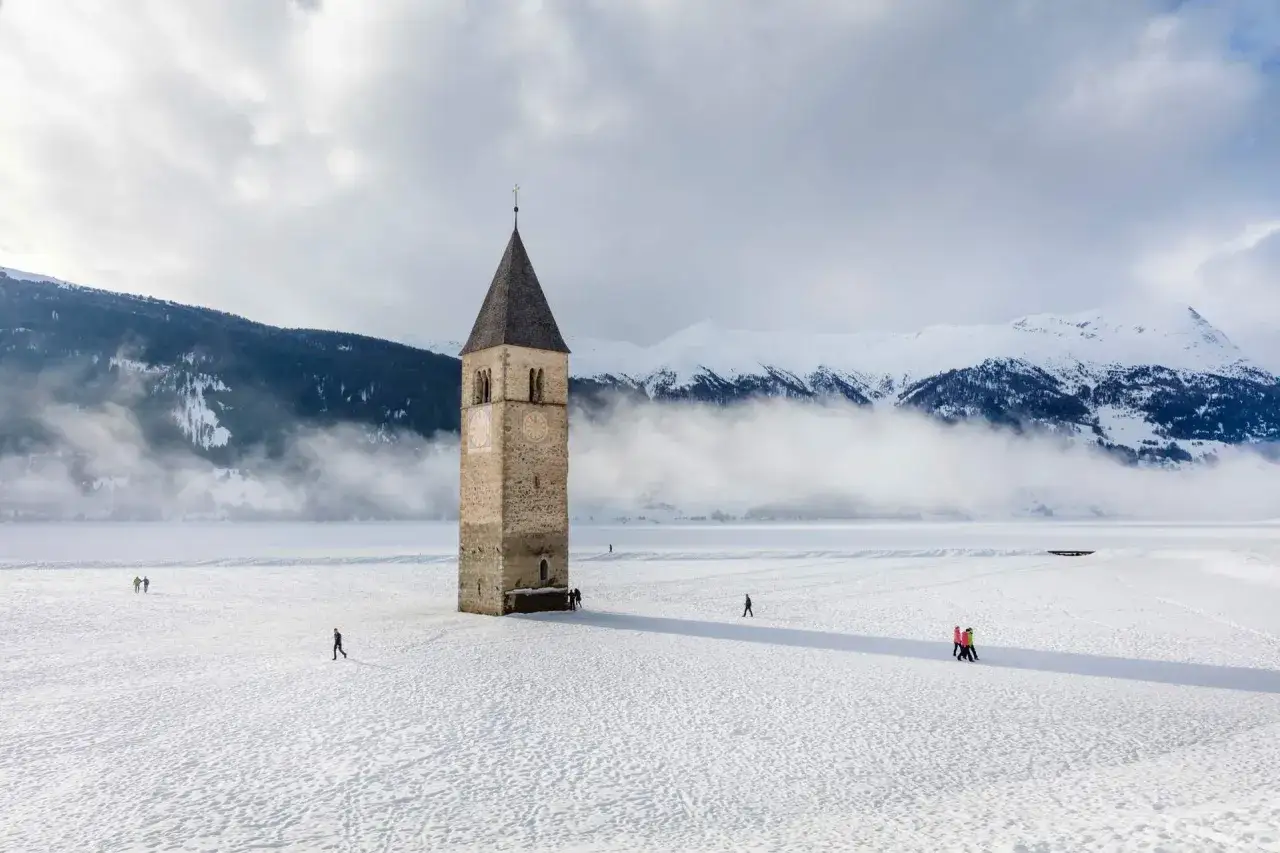 Un antico campanile nel lago ghiacciato, circondato da montagne innevate e nuvole basse. Alcune persone passeggiano sulla distesa bianca.
