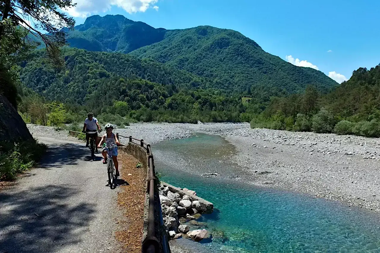 Ciclovia del Tagliamento: due ciclisti esplorano il sentiero panoramico lungo il fiume, circondati da montagne verdi e un cielo azzurro.