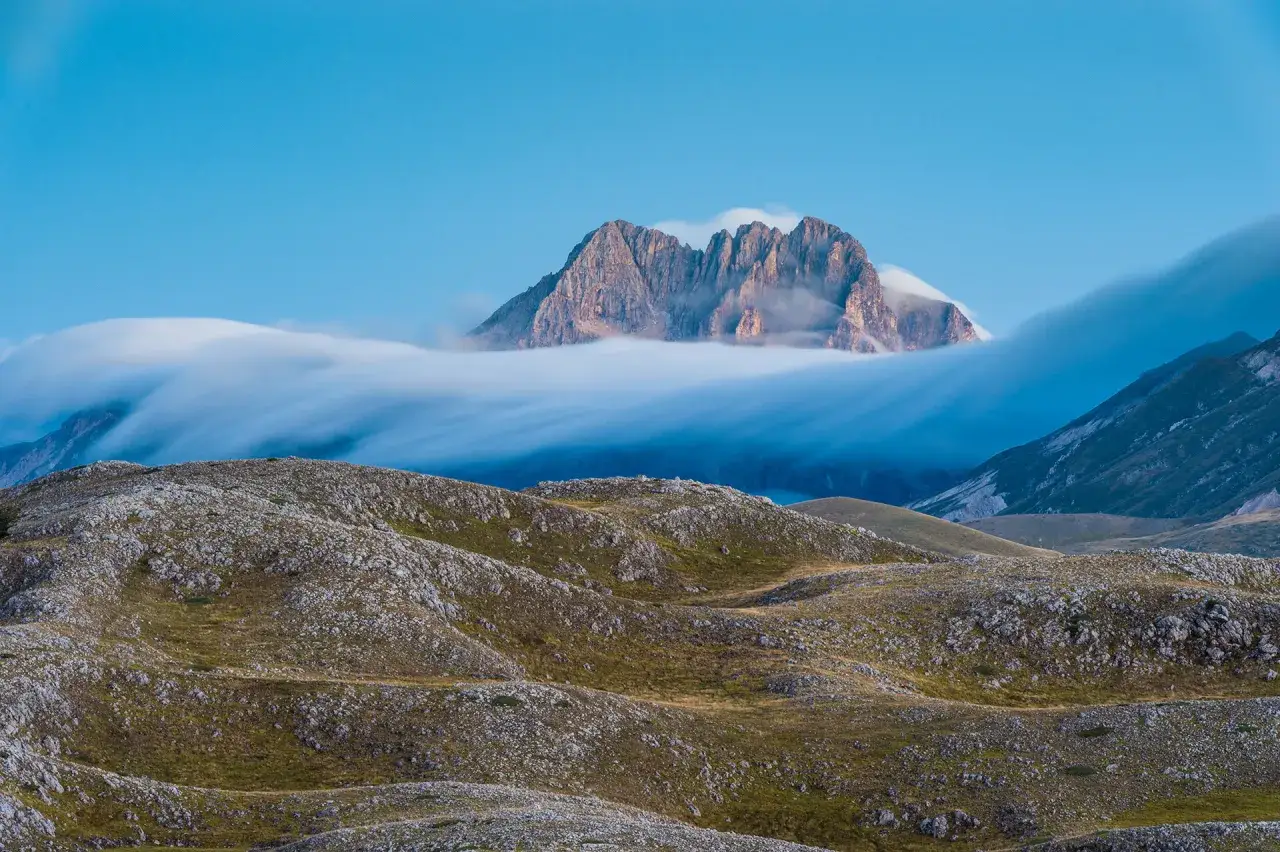 Vetta maestosa di una montagna del centro Italia emerge da un mare di nuvole che scorrono lente.