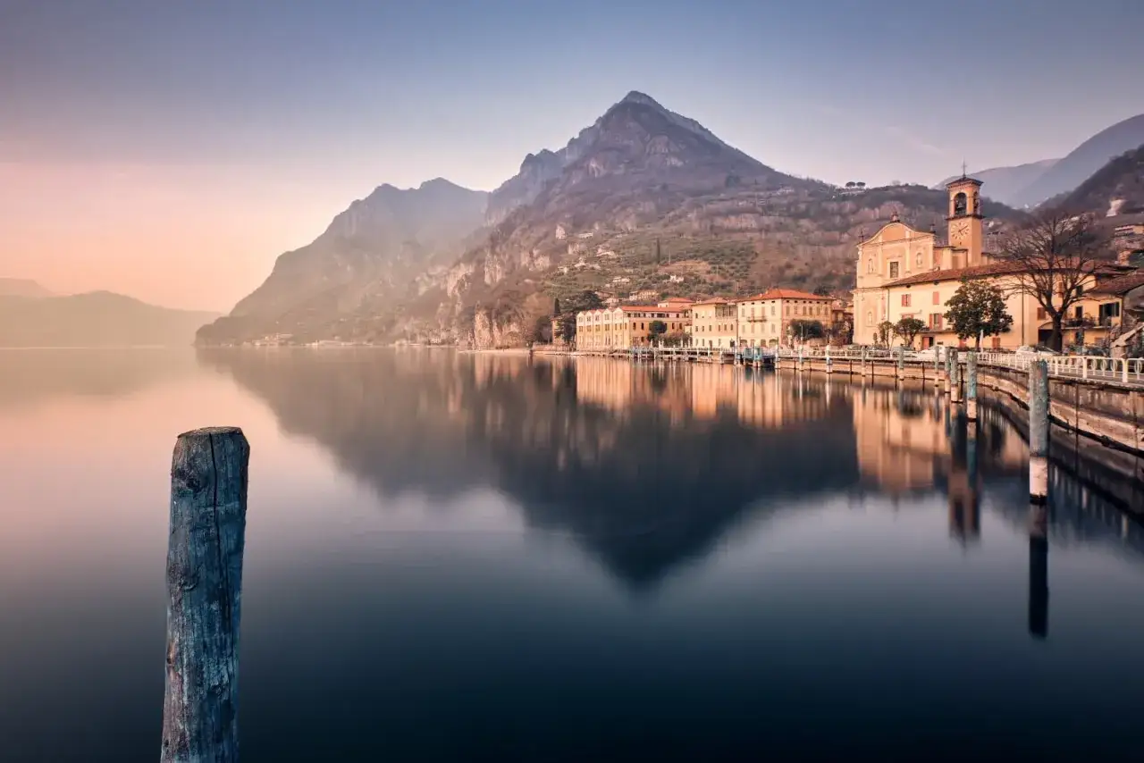 Un tranquillo lago vicino Milano, con montagne sullo sfondo e un villaggio pittoresco riflesso sull'acqua calma.