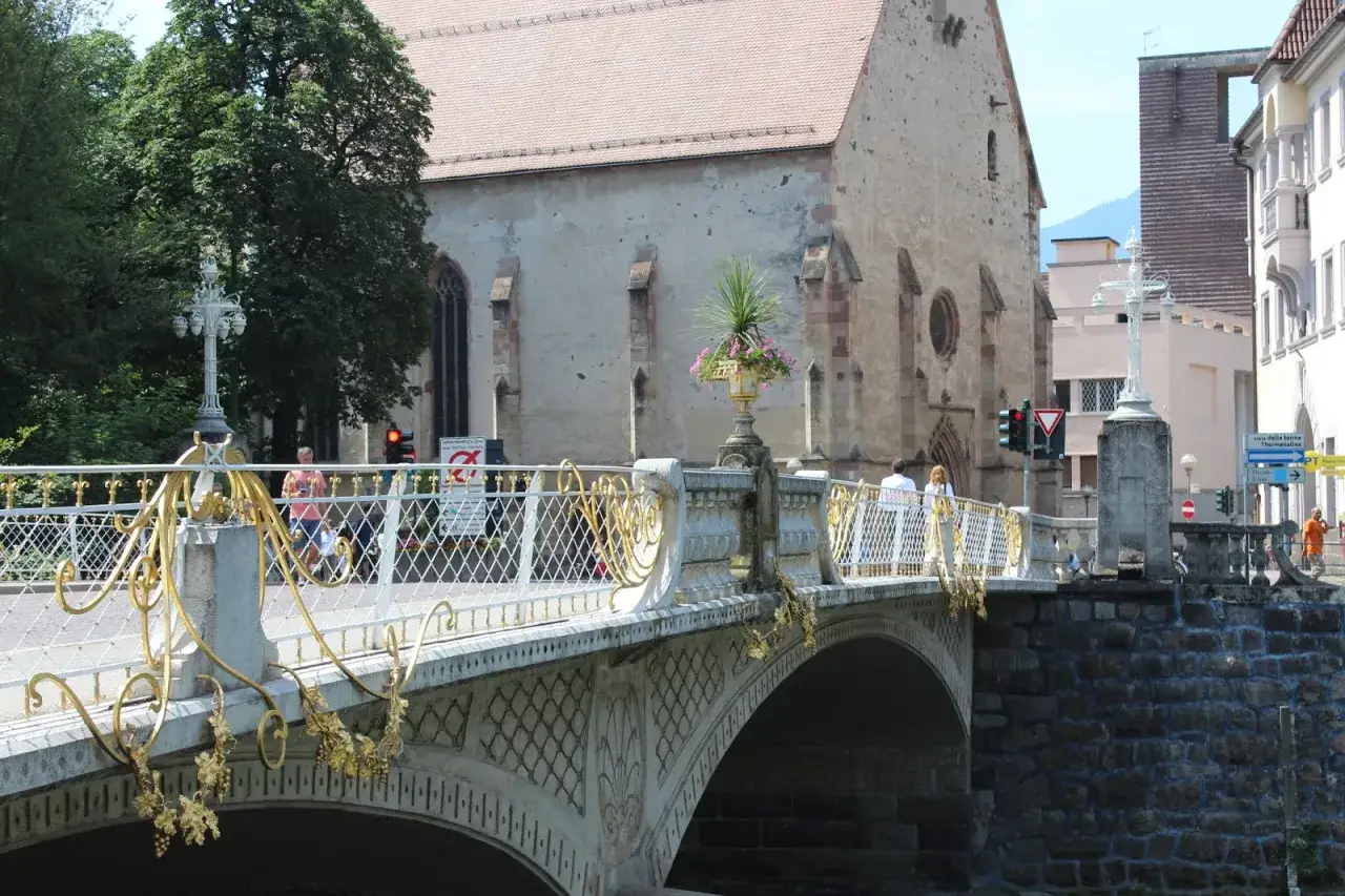Ponte decorato nel centro storico di Merano, con una chiesa sullo sfondo e persone che passeggiano.