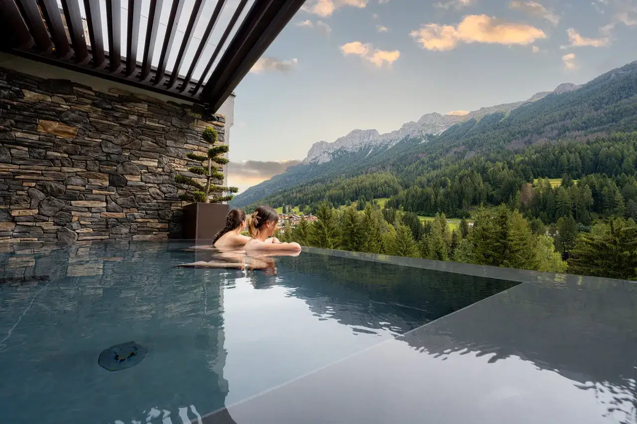 Due donne si rilassano in una piscina a sfioro con vista sulle montagne e foreste della Val di Fassa, un'esperienza rigenerante alle terme.