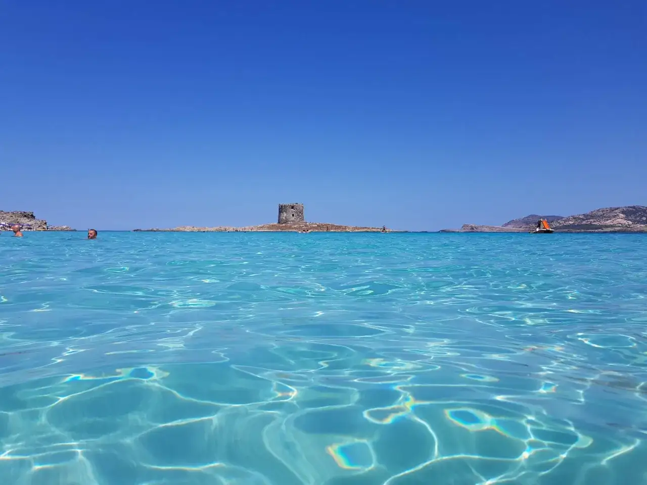 Acque cristalline del mare nord Italia, con una torre antica su un isolotto e una barca a vela arancione in lontananza.