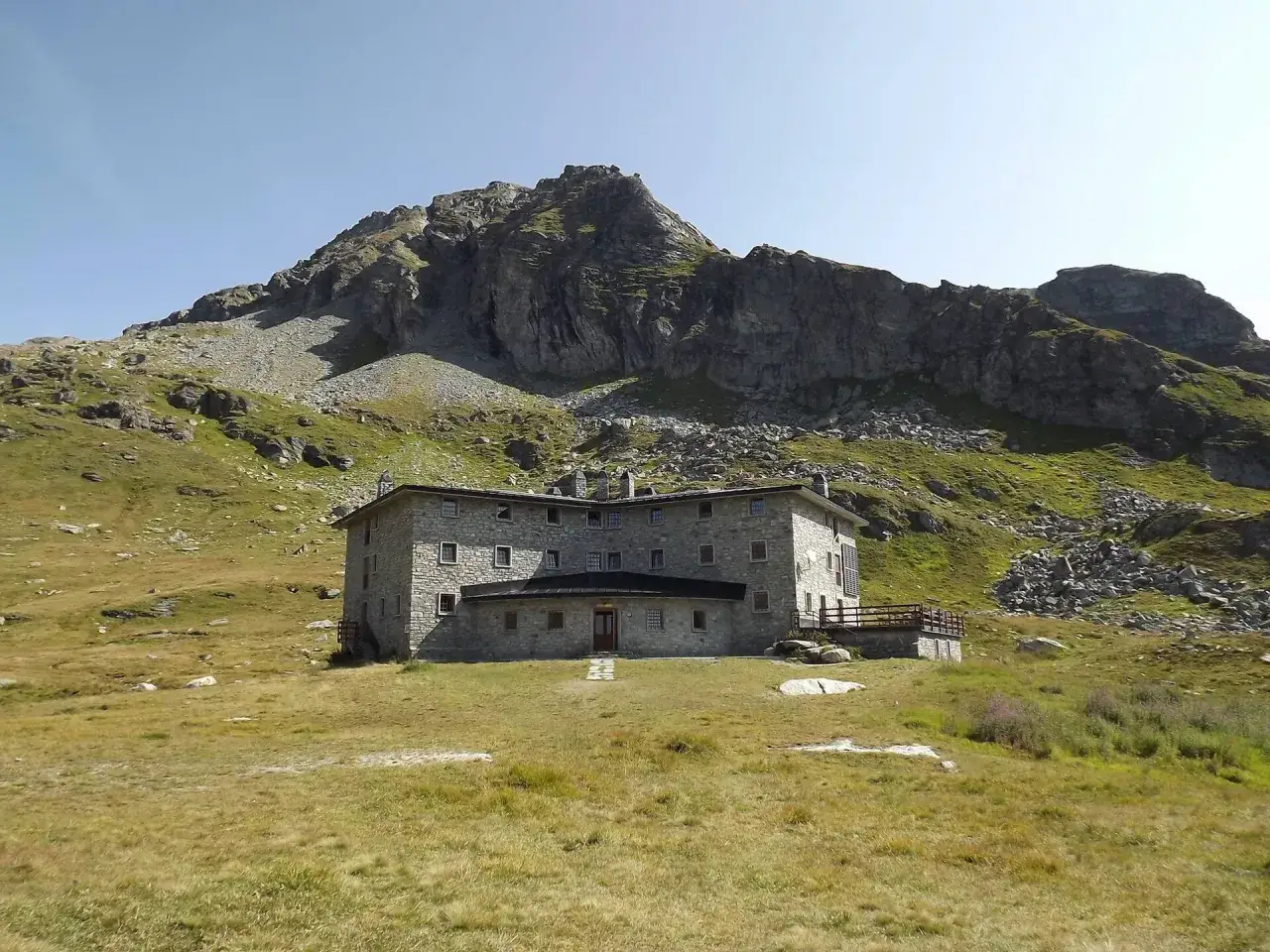 Rifugio in pietra con tetto scuro, circondato da prati verdi e rocce, sotto un imponente monte. Un luogo ideale per scoprire i rifugi Valle d'Aosta facili.