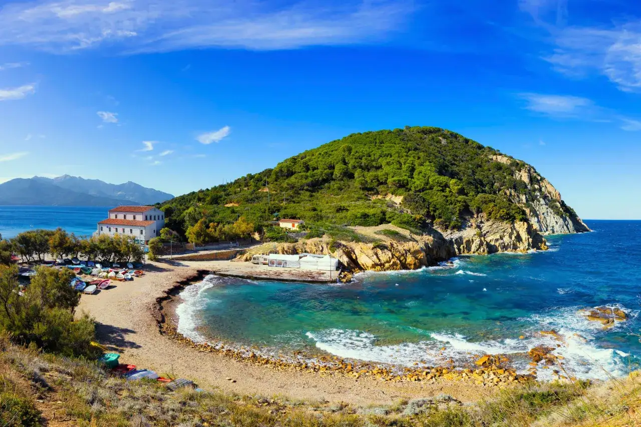 Baia con acque cristalline e una collina verdeggiante sull'isola d'Elba. Barche a riva e un edificio bianco completano la scena.
