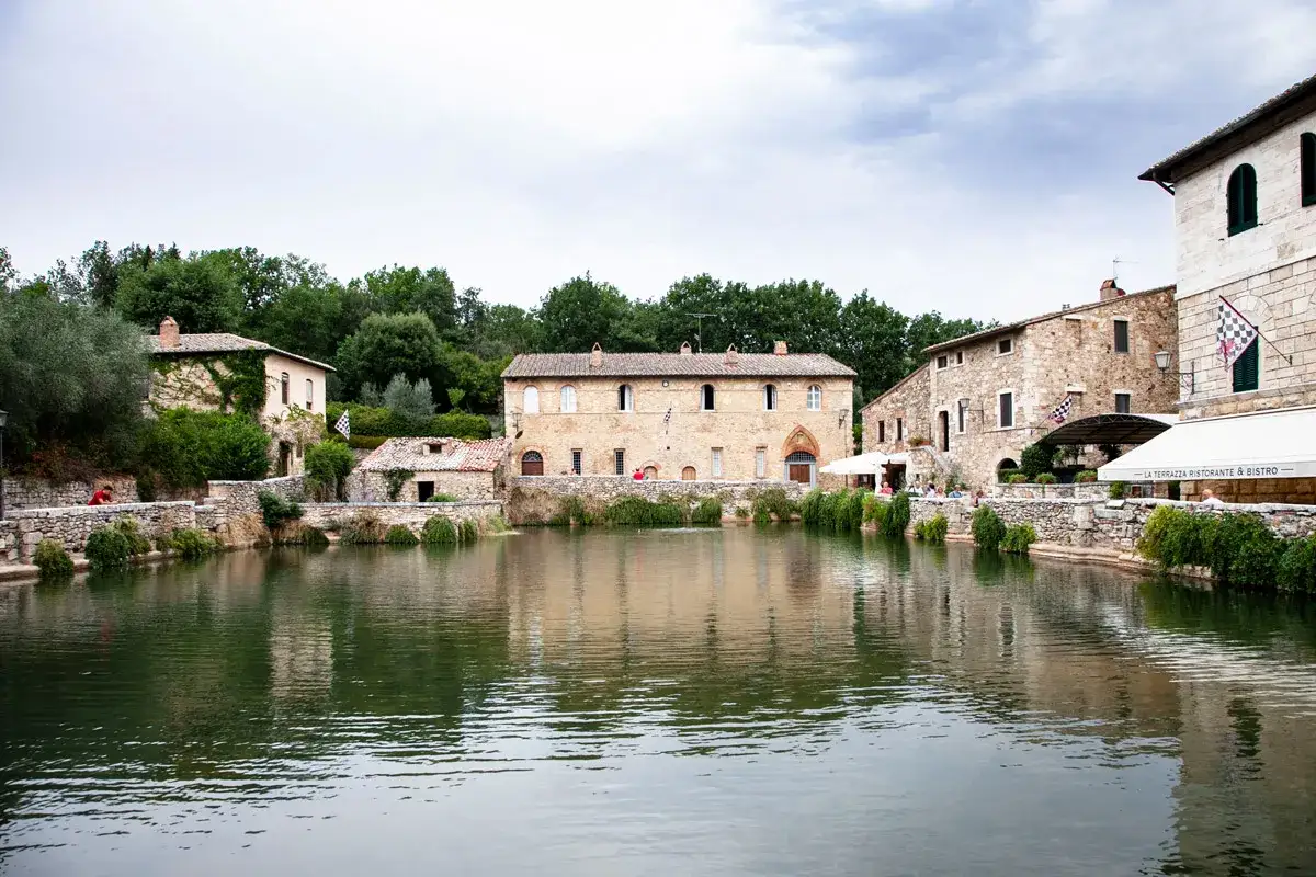 Antiche terme nel centro Italia, con edifici in pietra che si affacciano su uno specchio d'acqua tranquillo.
