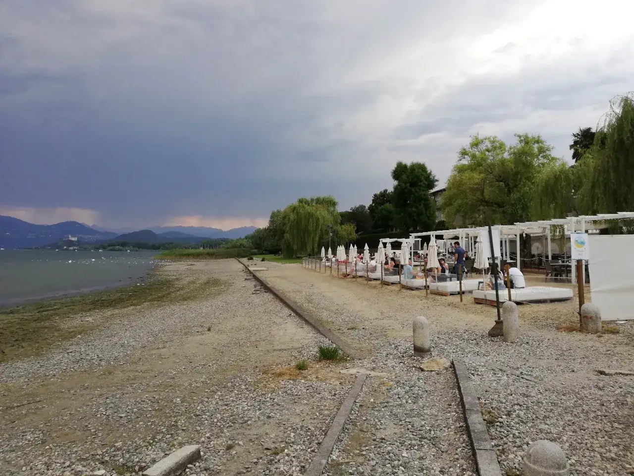 Lido di Lisanza con lettini bianchi e ombrelloni, gente rilassata, lago e montagne sullo sfondo.