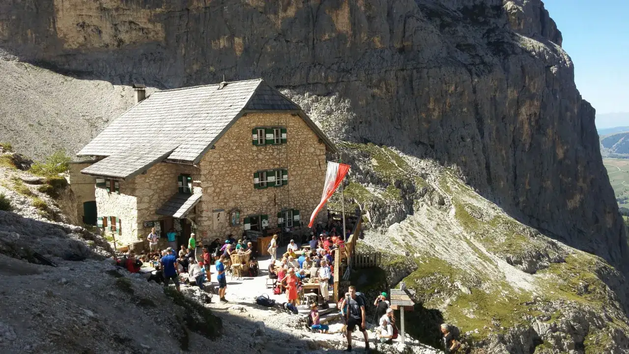 Rifugio alpino affollato, meta di escursionisti per attività in montagna. Persone sedute ai tavoli all'aperto, godendosi il sole e il panorama.