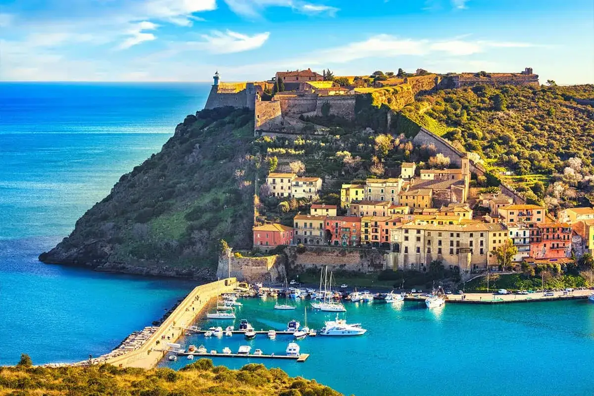 Incantevole località mare toscana con un borgo colorato arroccato su una collina, un porto con barche e un'antica fortezza con faro.