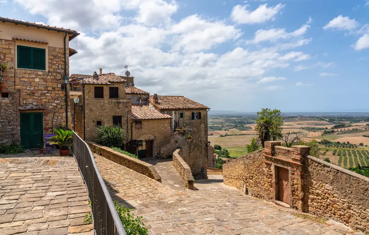 Un borgo antico in pietra si affaccia sulla campagna toscana lungo il mare, con case in mattoni e tetti rossi che scendono verso la costa.