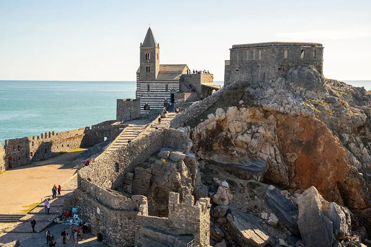 Castello e chiesa a picco sul mare, un gioiello della riviera di Levante. Turisti esplorano antiche mura e sentieri rocciosi.