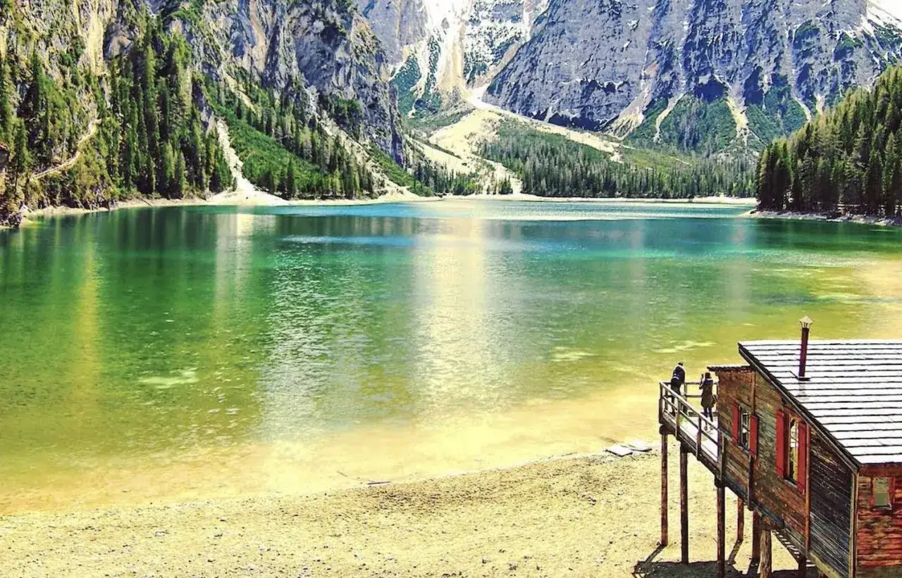 Lago alpino dalle acque smeraldine, con un rifugio in legno e due persone che ammirano il panorama. Un'idea per un itinerario Trentino Alto Adige.