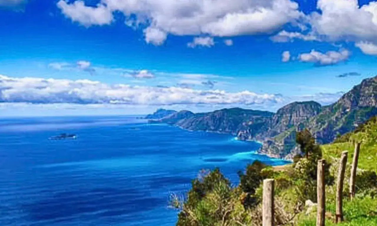 Sentiero costiera amalfitana con mare blu intenso, montagne scoscese e cielo azzurro con nuvole bianche.
