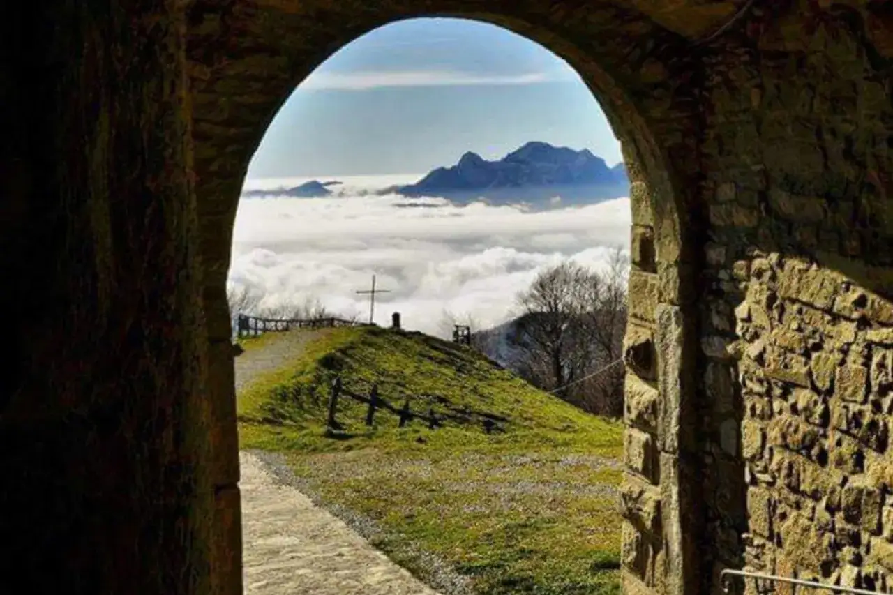 Un sentiero sale verso un crocifisso, sopra un mare di nuvole, con montagne sullo sfondo. Un'antica porta in pietra incornicia la vista, come un invito a percorrere la via del volto santo.