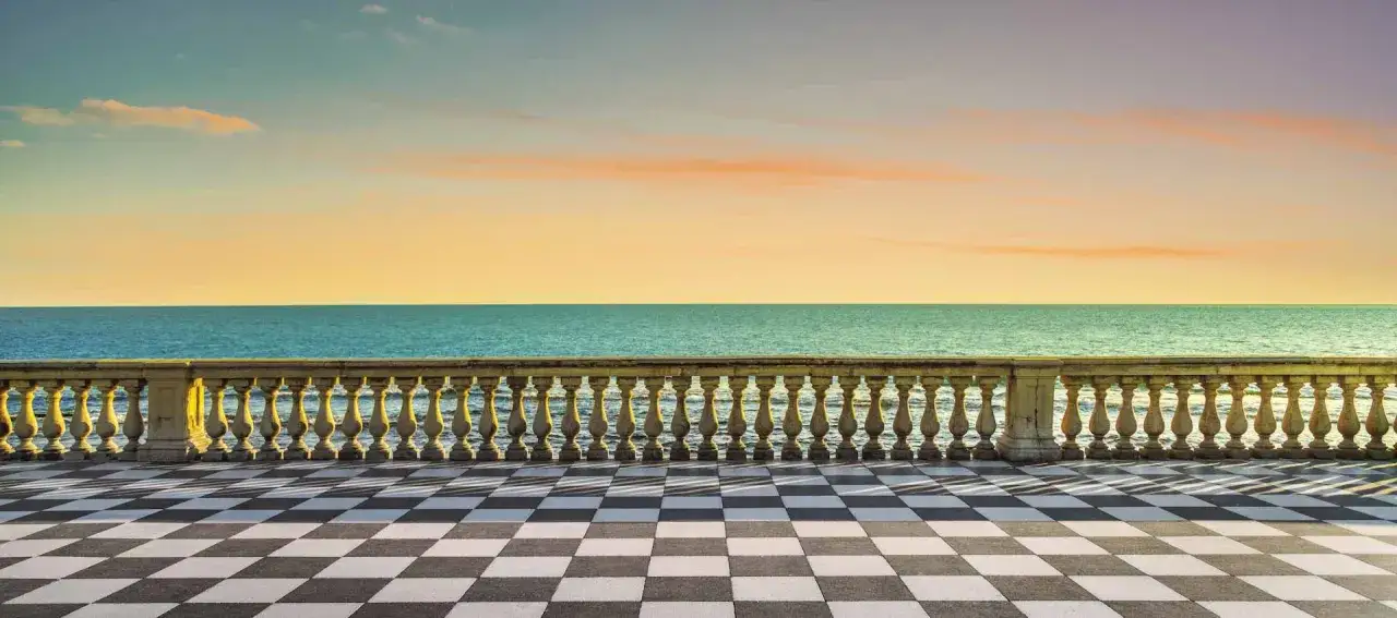 Terrazza con pavimento a scacchi e balaustra in pietra si affaccia sul mare calmo al tramonto, ideale per una passeggiata al mare.