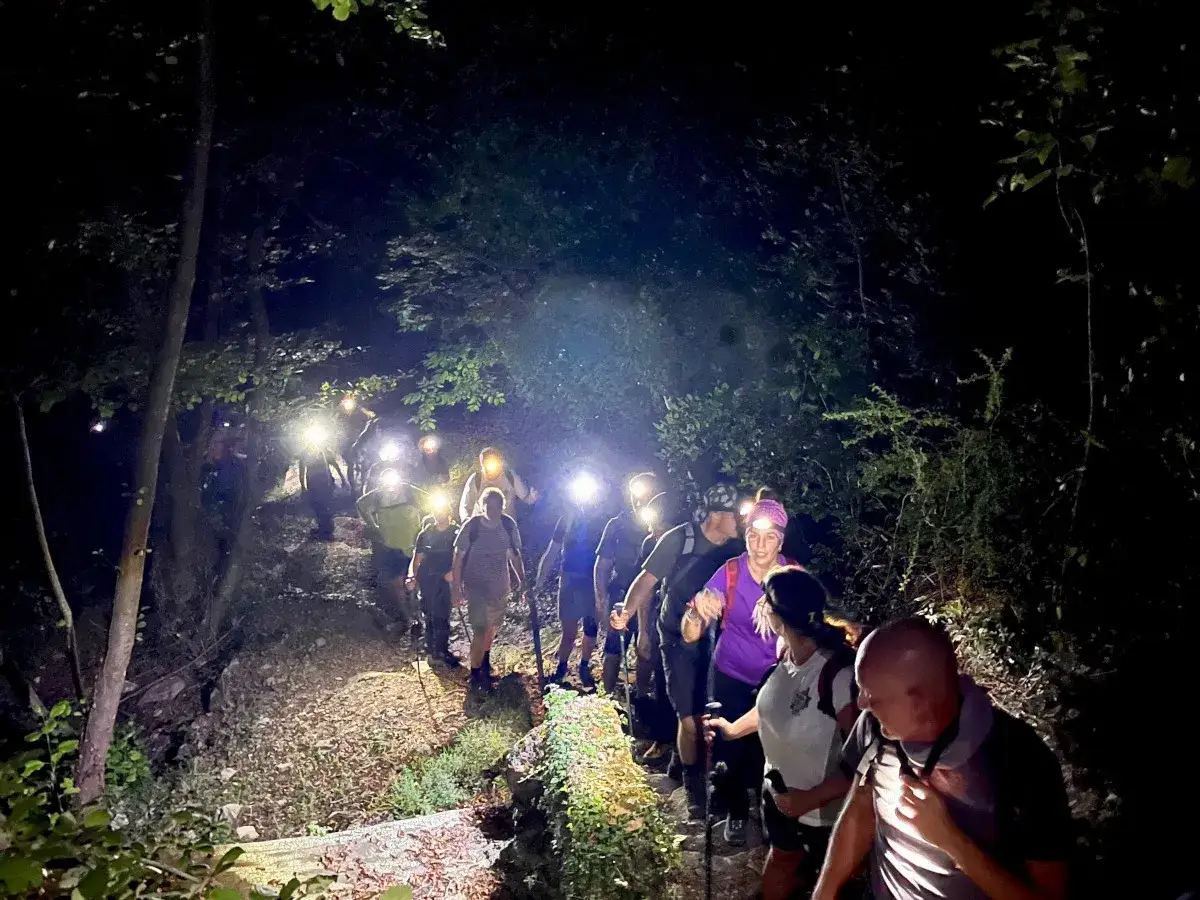 Un gruppo di escursionisti con luci frontali illumina un sentiero nella montagna di notte, tra alberi e vegetazione.