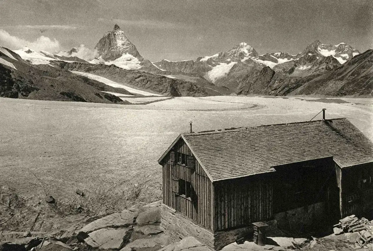 Rifugio Mantova Vioz, un piccolo edificio in legno, si erge su rocce con un vasto ghiacciaio e montagne innevate sullo sfondo.