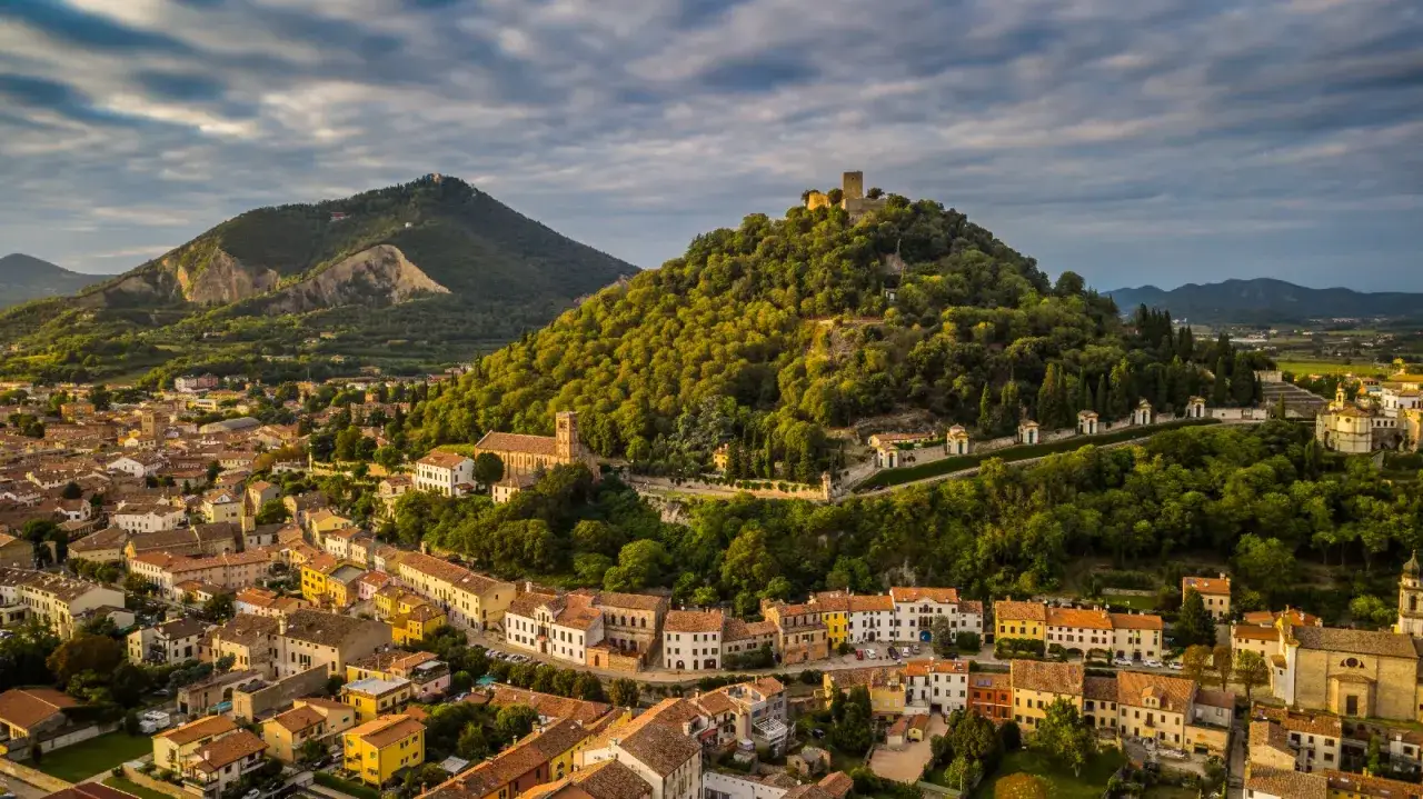 Panoramica aerea di borghi medievali veneti con castello in cima a una collina verdeggiante, case colorate e chiese antiche.