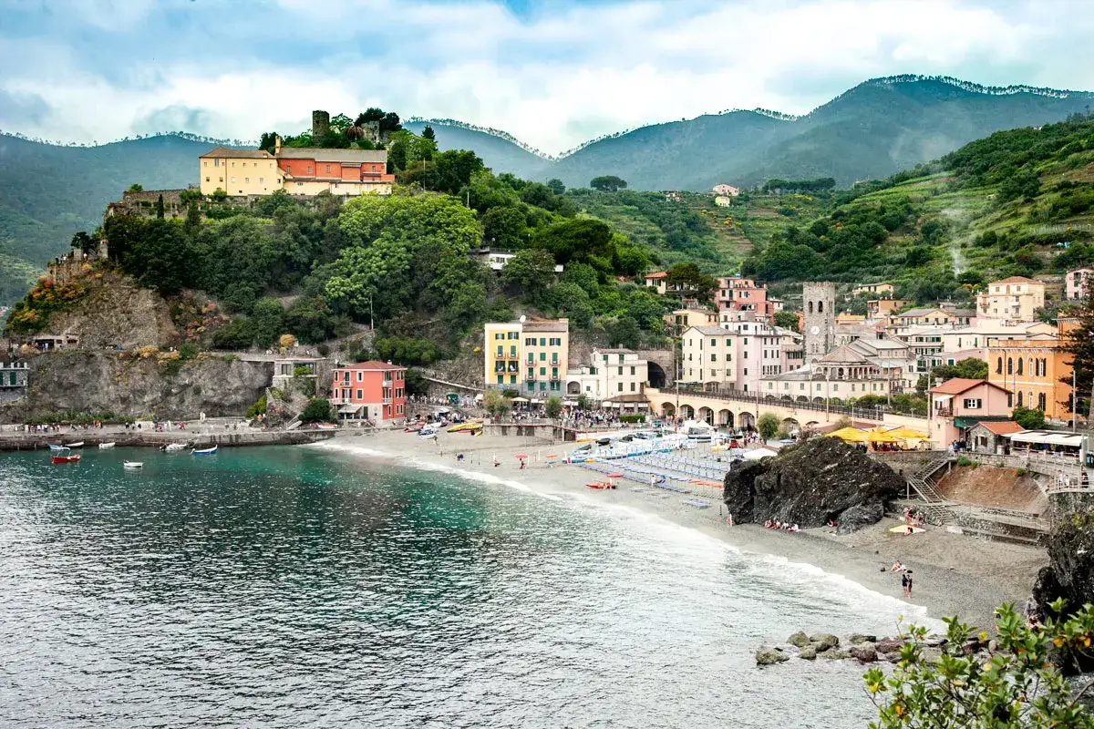Colorate case a picco sul mare, una spiaggia affollata e montagne verdi sullo sfondo, un tipico scorcio delle Cinque Terre.