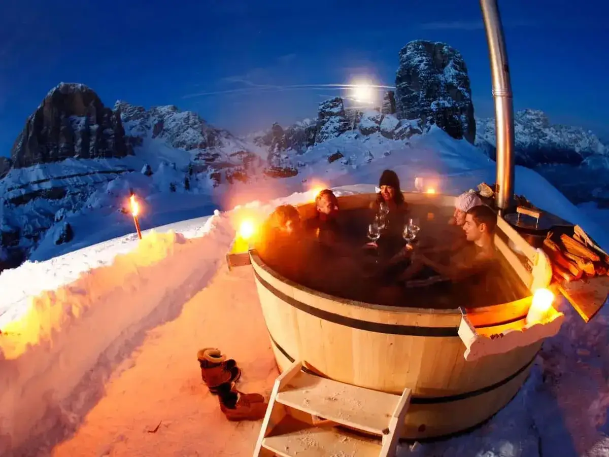 Un gruppo di amici si rilassa in una vasca idromassaggio all'aperto, circondata dalla neve e dalle maestose Dolomiti. Un'esperienza indimenticabile tra i migliori rifugi dove dormire Dolomiti.