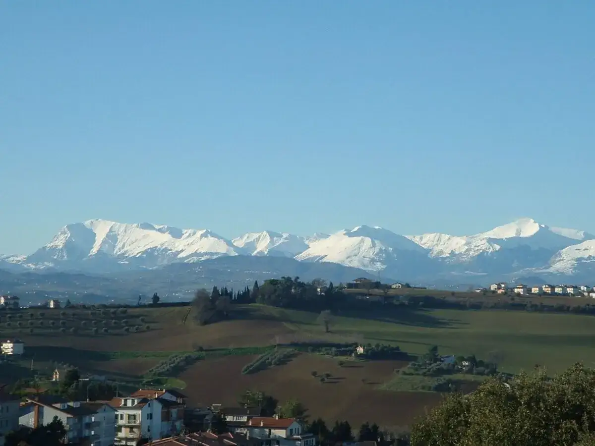 Paesaggio innevato con montagne maestose sullo sfondo, colline verdi e case sparse. Un'incantevole vista di San Ginesio, Marche.