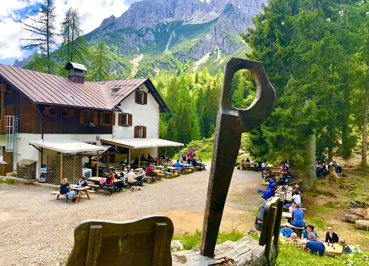 Rifugio alpino con tavoli all'aperto, persone che mangiano e bevono, circondato da alberi e montagne imponenti. I forni di sopra sono un luogo ideale per una pausa.