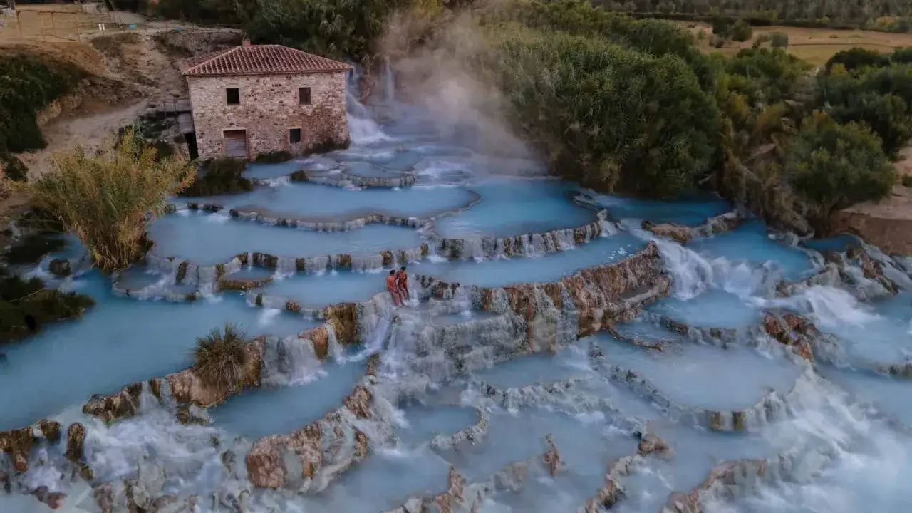 Cascate naturali con acqua azzurra e vapore, un antico casale sullo sfondo. Un luogo magico per godersi le terme di Saturnia gratis.