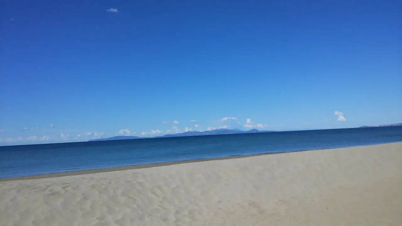 Spiaggia dorata e mare calmo a Marina di Follonica, con montagne all'orizzonte sotto un cielo azzurro limpido.